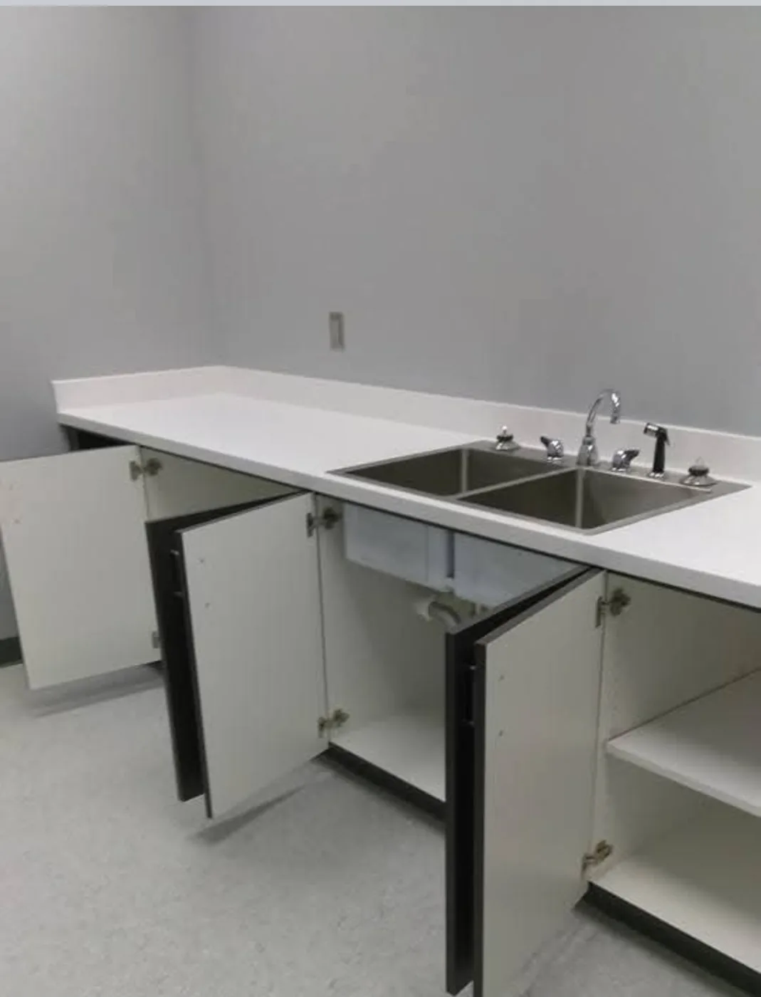A commercial break-room with a long white counter, a stainless steel double sink and clean white cabinets with the doors open, freshly cleaned