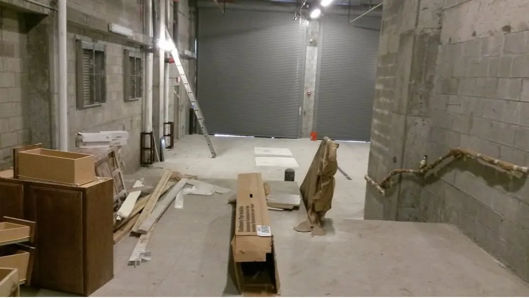 An active commercial construction interior with leftover lumber, cardboard, a ladder and wrapped materials on a bare concrete floor, before the post-construction clean