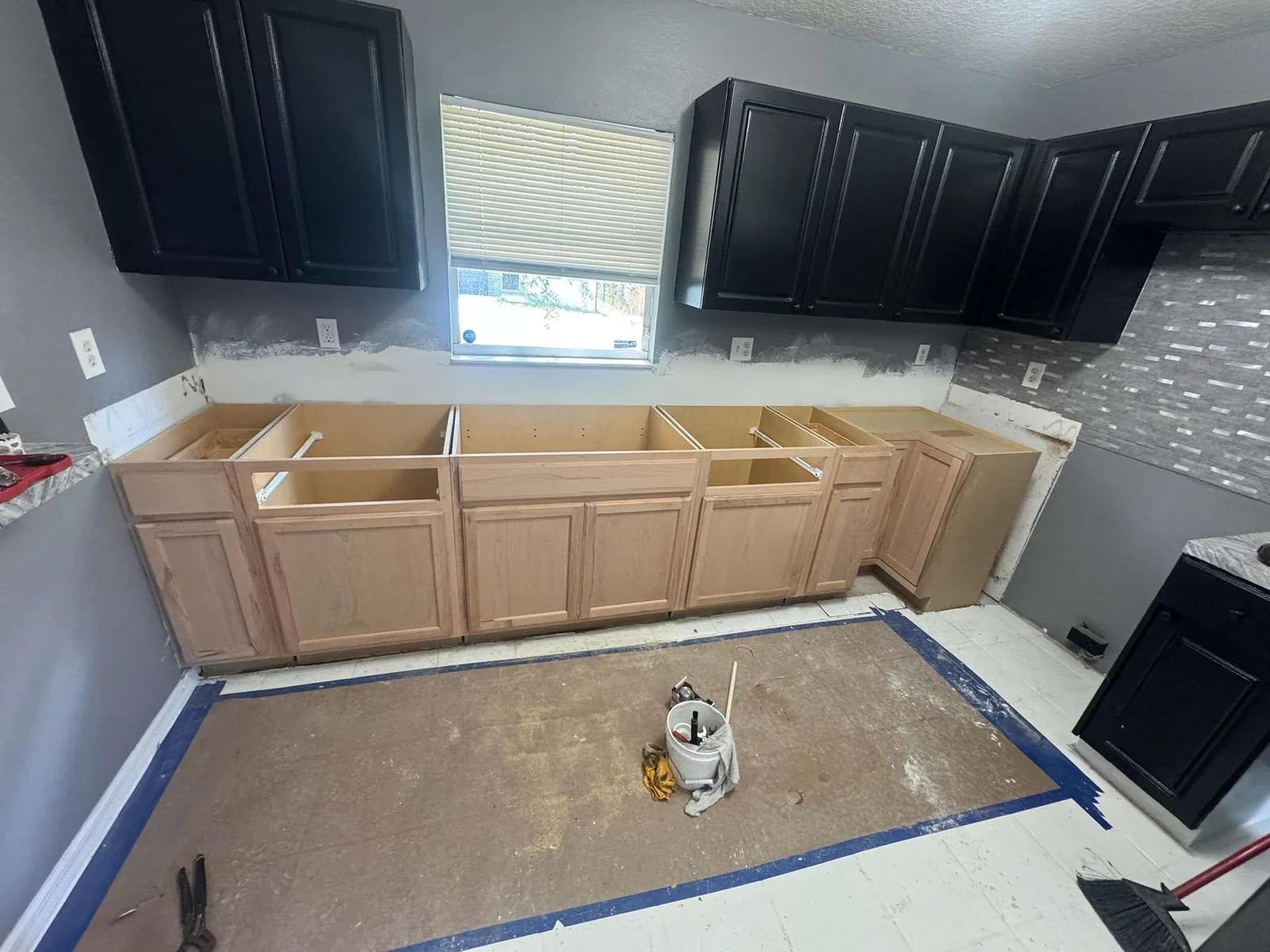 A wide view of the same Tampa kitchen mid-rehabilitation, black upper cabinets painted but the raw new base cabinets set with no countertop yet and the floor stripped down and taped off