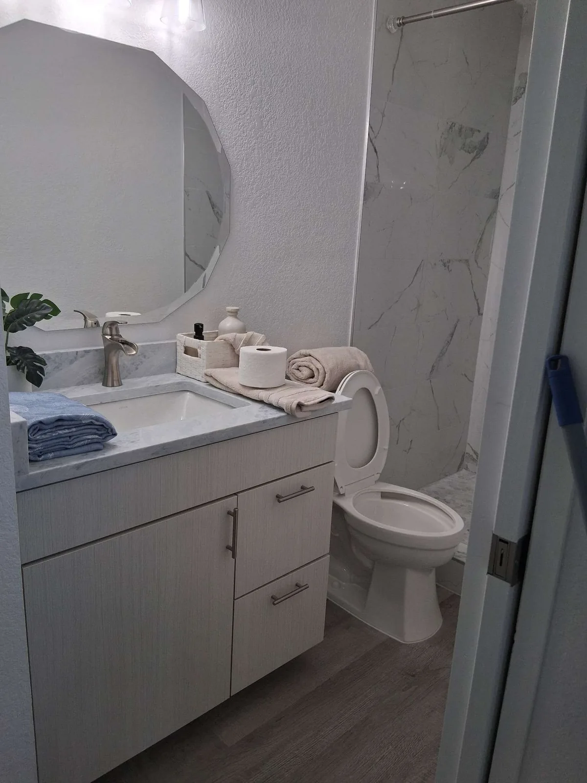 A furnished-rental bathroom with a marble-top vanity, brushed-nickel faucet, octagonal mirror, and folded towels and toiletries staged on the counter, with a marble walk-in shower visible behind