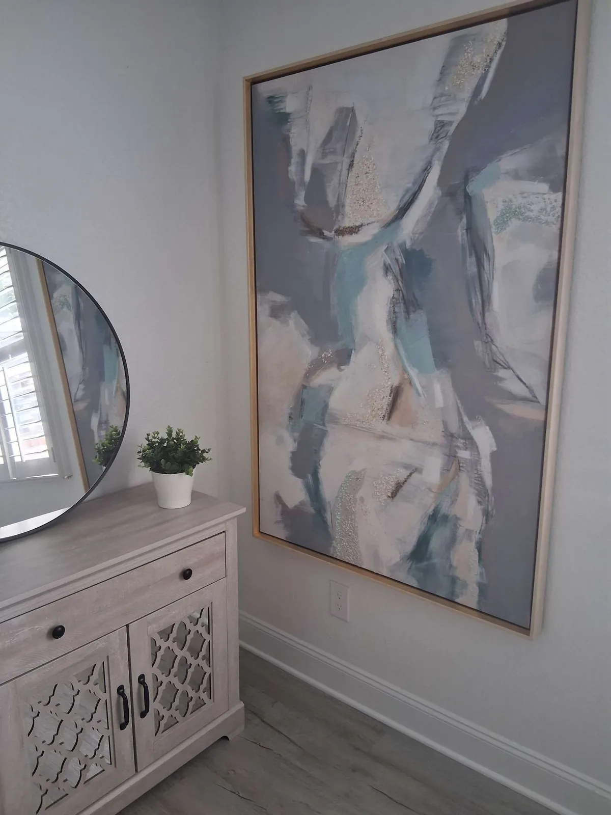 A furnished-rental entryway with a round black-framed mirror and a small potted plant on a light wood console table, and a large framed abstract painting on the adjacent wall