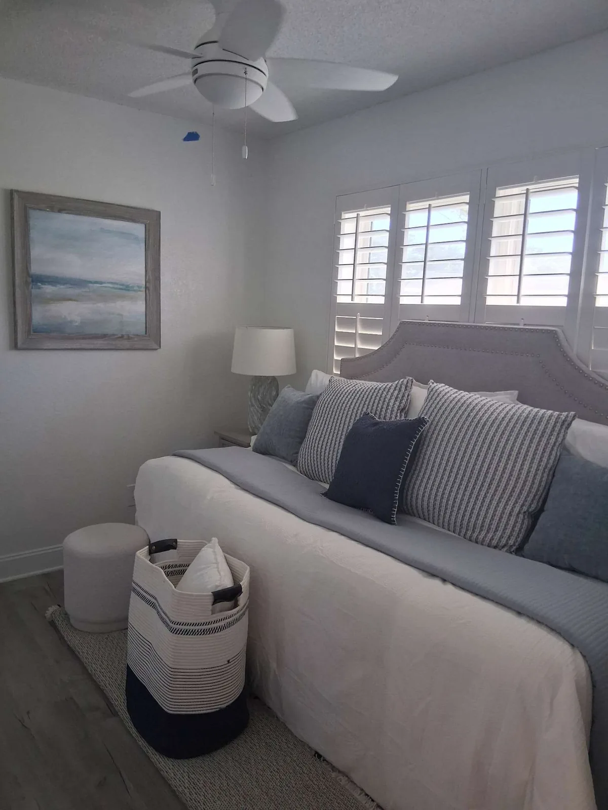A furnished-rental guest bedroom with a made bed, layered blue and grey striped pillows against an upholstered headboard, a ceiling fan overhead, plantation shutters, and a framed coastal painting on the wall