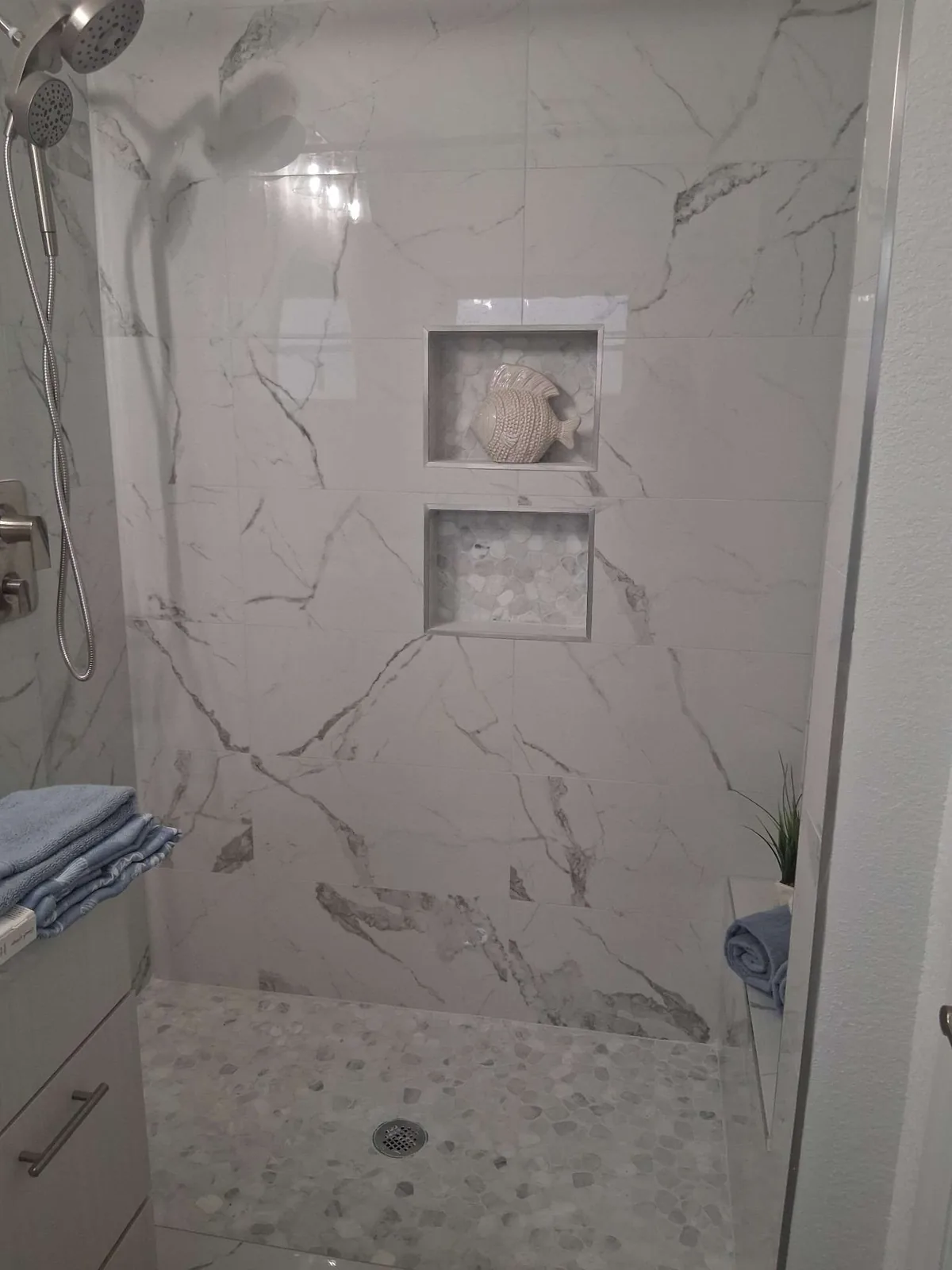 A marble-tiled walk-in shower with two tile niches, a pebble-mosaic floor, and folded towels on the bench and vanity, in a furnished-rental bathroom